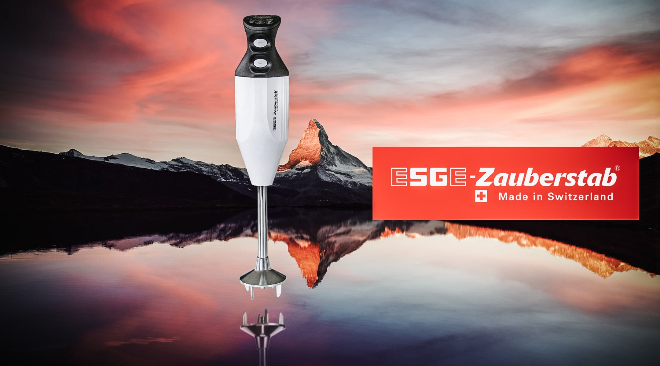 Ein weißer ESGE-Zauberstab-Stabmixer steht vor einer malerischen Berglandschaft mit einem spiegelnden See und rosa Himmel, neben einem roten ESGE-Zauberstab-Logo „Made in Switzerland“.