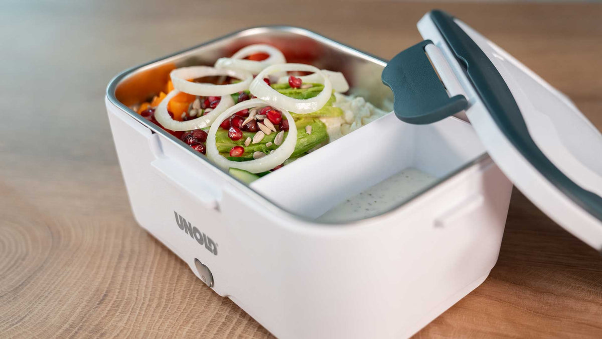 Eine weiße Lunchbox mit Metallfach, in dem sich ein Salat mit Zwiebelringen, Granatapfelkernen und Gemüse befindet. Der Deckel ist teilweise geöffnet und gibt den Blick auf ein separates Fach mit einem cremigen Dip frei.