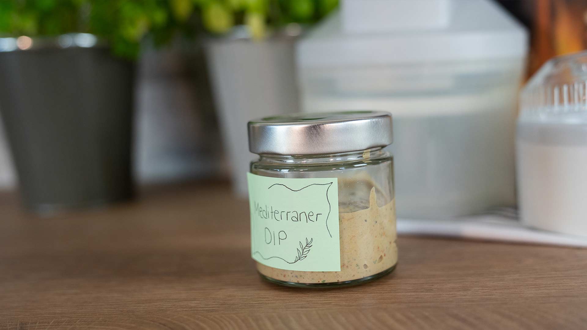 Ein Glasgefäß mit silbernem Deckel und der Aufschrift „Mediterraner Dip“, das einen hellbraunen Brotaufstrich enthält, steht auf einer Holzoberfläche, im Hintergrund sind unscharfe Küchenutensilien zu sehen.