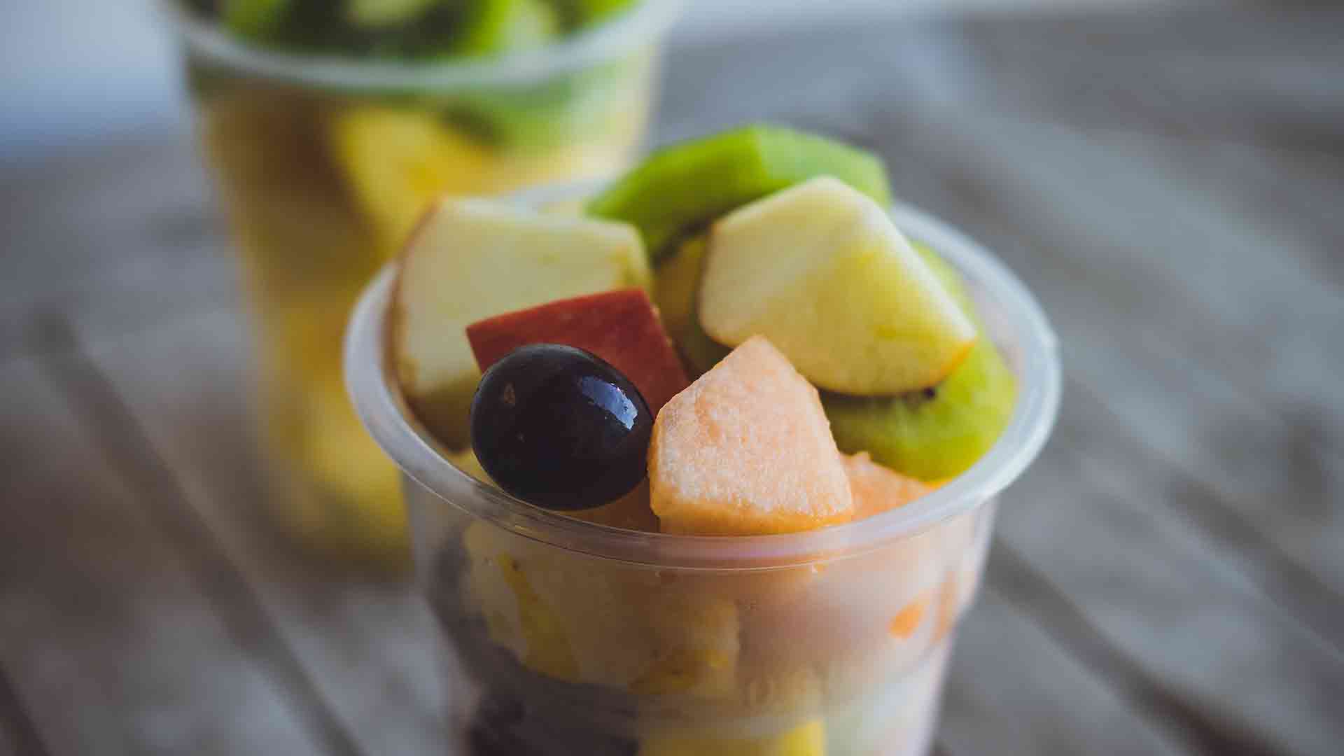Ein durchsichtiger Plastikbecher, gefüllt mit verschiedenen frischen Fruchtstücken, darunter Apfel, Traube, Melone und Kiwi, steht auf einer Holzoberfläche, während im Hintergrund ein weiterer Becher mit Obst leicht unscharf zu sehen ist.
