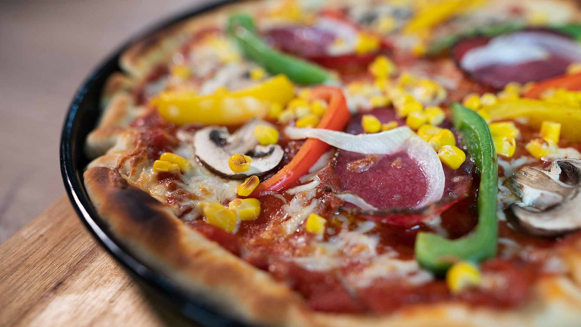 Nahaufnahme einer Pizza mit Peperoni, gelbem Mais, geschnittenen Champignons, grünen und gelben Paprika, Zwiebeln und geschmolzenem Käse auf einer Tomatensoßenbasis.