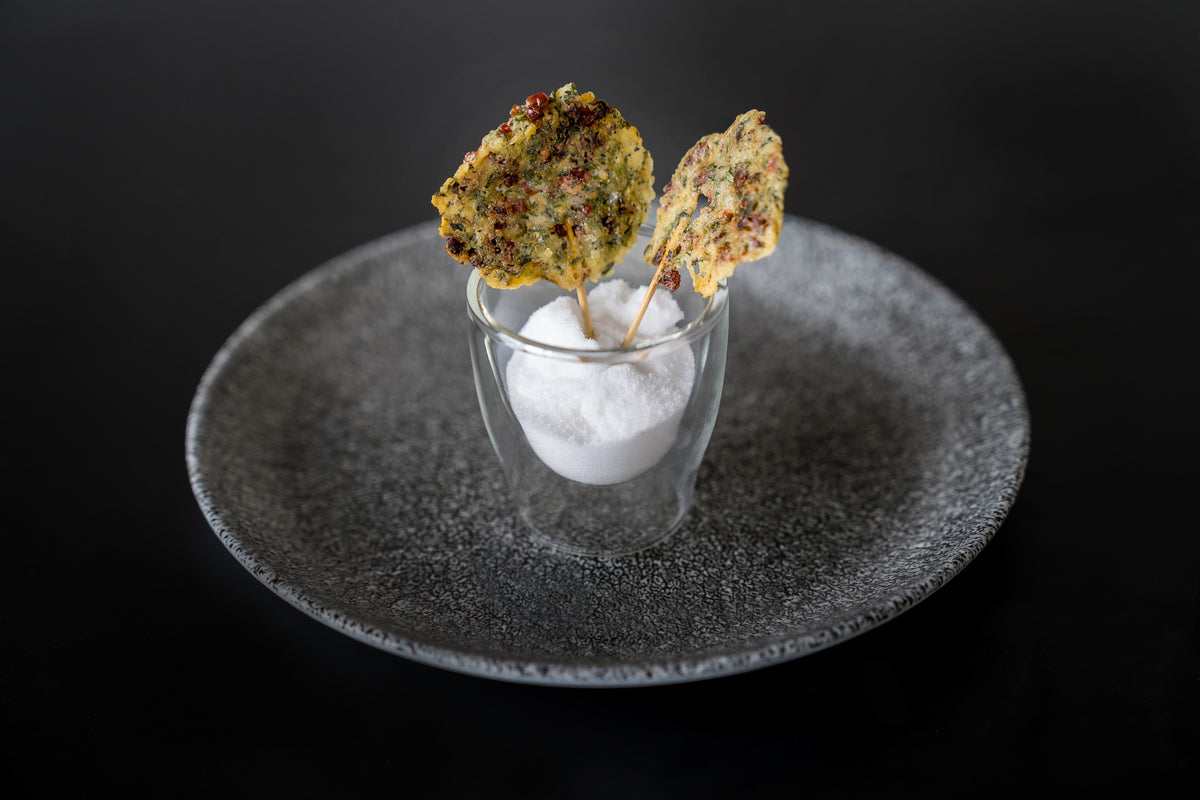 Auf einem grau strukturierten Teller liegen zwei blattförmige, gewürzte Cracker am Stiel und ein Glas mit weißem Salz, fotografiert mit dem Raclette-Rezeptbuch Deluxe von Unold vor schwarzem Hintergrund.