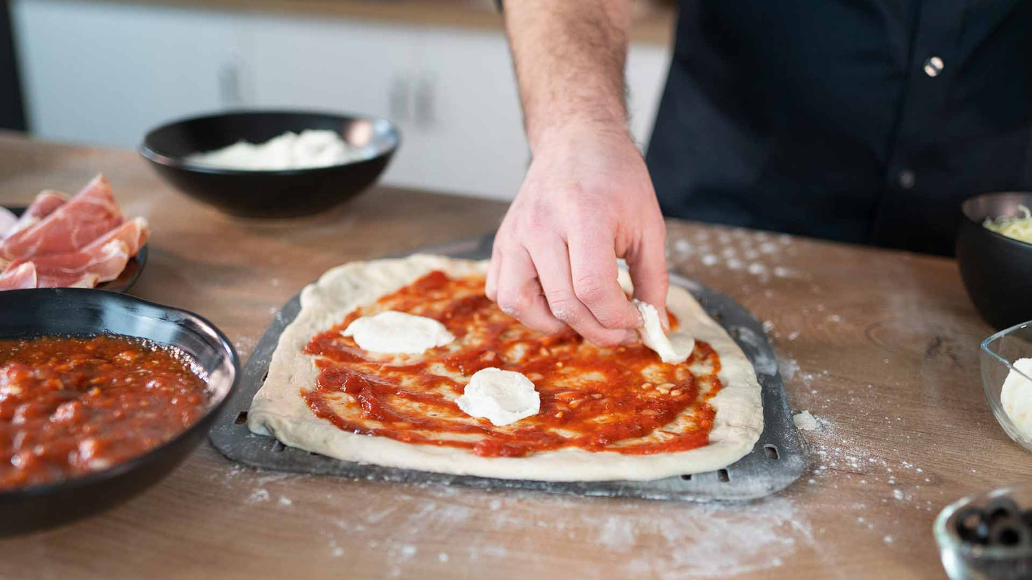 Eine Person legt Käsestücke auf einen mit Tomatensoße belegten Pizzateig auf einem Backblech. Daneben stehen Schüsseln mit Zutaten auf einer Holzarbeitsplatte.