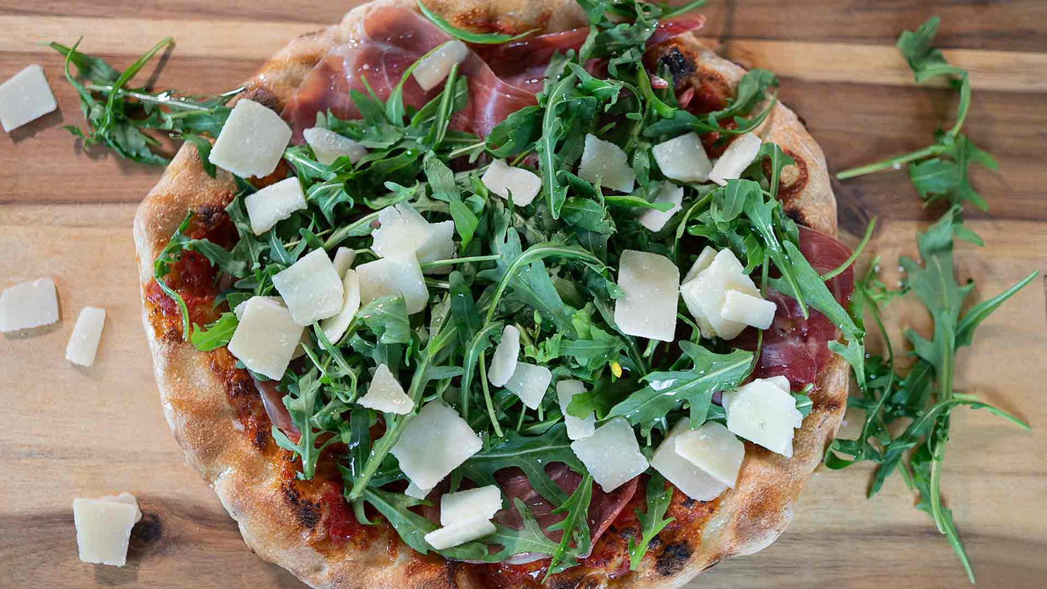 Eine Pizza mit Rucola, dünnen Schinkenscheiben und gehobeltem Parmesan, präsentiert auf einer Holzoberfläche. Einige Rucola- und Käsestücke sind auf der Pizza verstreut.