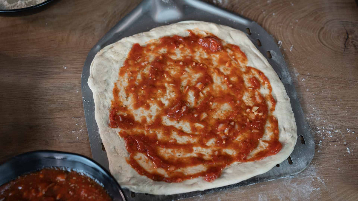 Ein roher Pizzateig auf einer Pizzaschaufel aus Metall mit darauf verteilter Tomatensoße, bereit für weiteren Belag, auf einer Holzoberfläche.