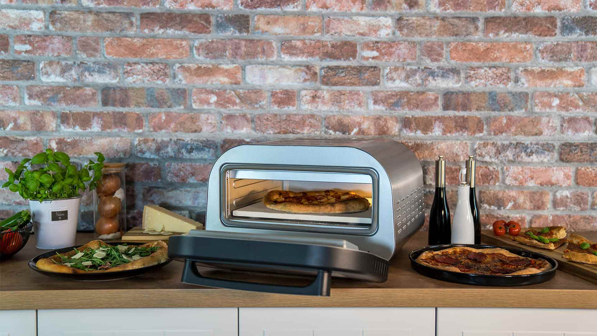 Auf einer Küchentheke steht ein Pizzaofen mit einer Pizza darin, umgeben von drei Tellern Pizza, einem Topf mit Basilikum, Käse und Gewürzen, vor dem Hintergrund einer Ziegelwand.