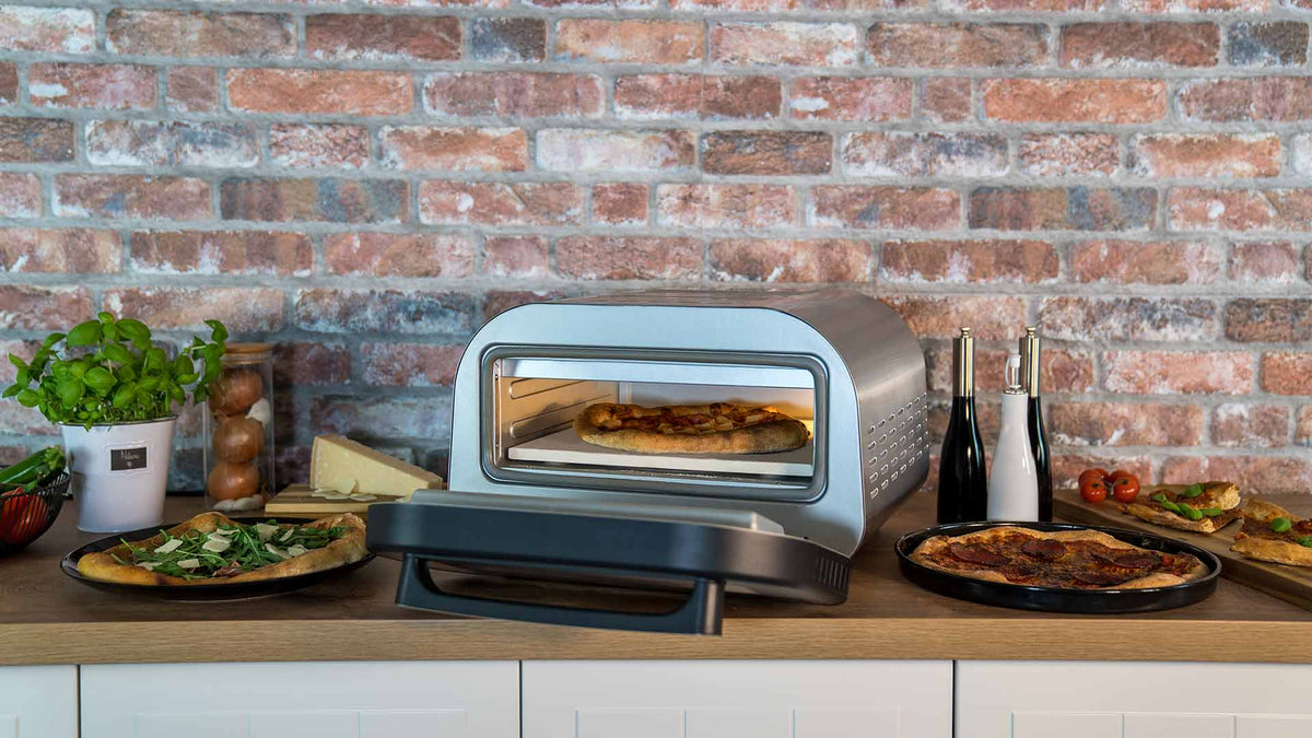 Auf einer Küchentheke steht ein Pizzaofen mit einer Pizza darin, umgeben von Tellern mit gebackener Pizza, Kräutern, Käse, Gemüse und Küchenutensilien vor dem Hintergrund einer Backsteinmauer.