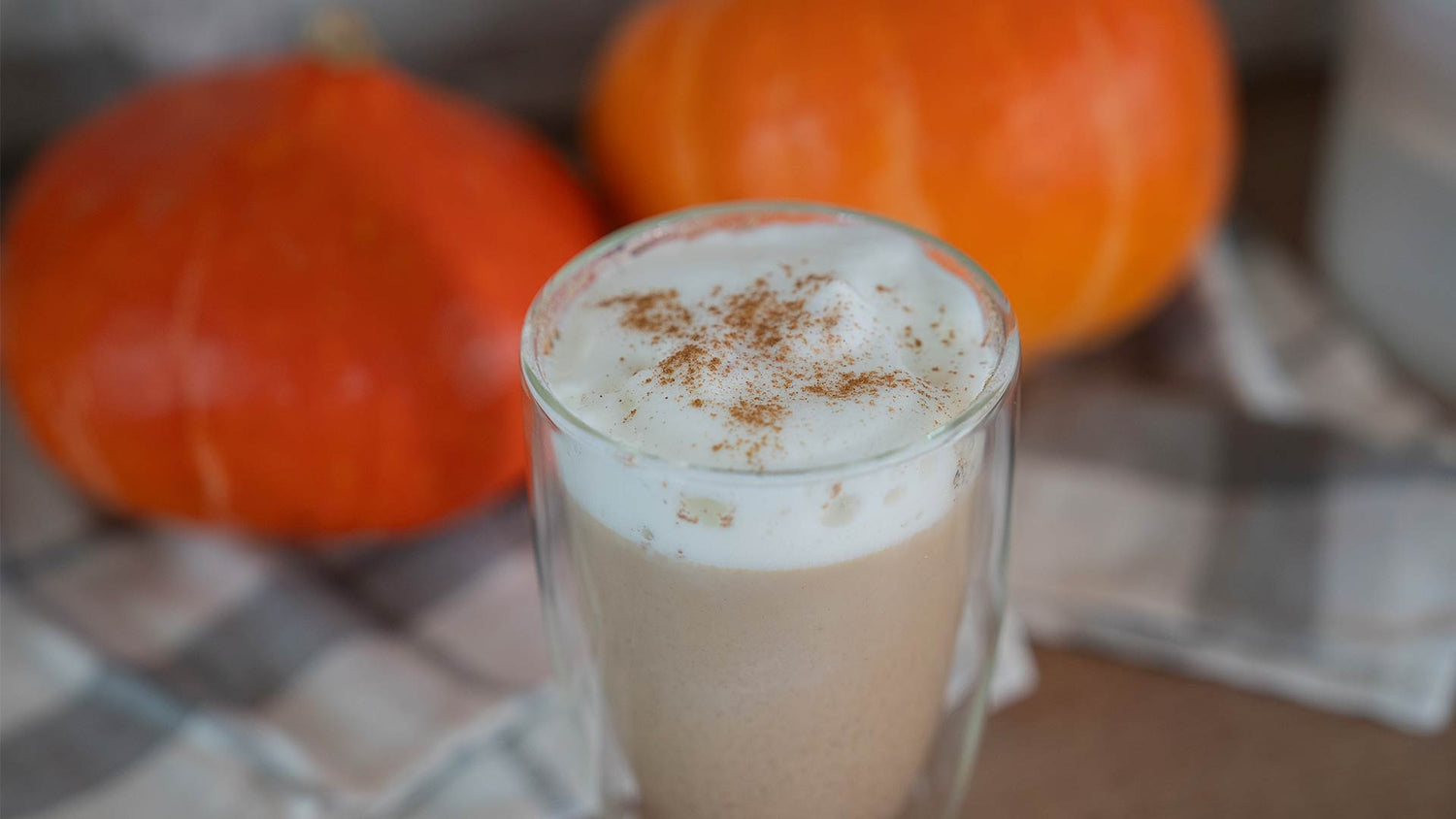 Ein Glas Pumpkin Spice Latte mit aufgeschäumter Milch und einer Prise Zimt steht auf einem karierten Tuch, im Hintergrund sind zwei kleine orangefarbene Kürbisse zu sehen.