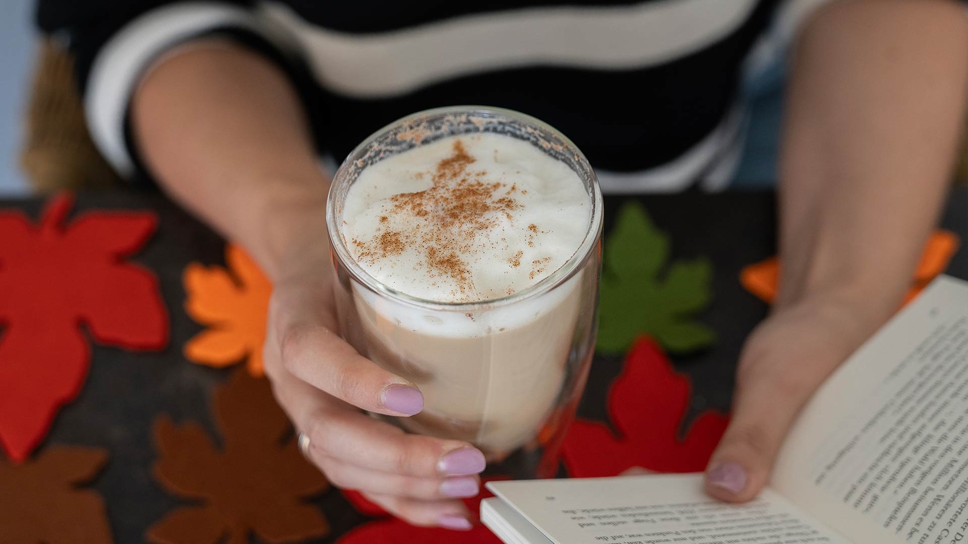 Eine Person hält ein Glas schaumigen Kaffee mit Zimt, umgeben von bunten Herbstblättern aus Filz, und liest dabei ein Buch.