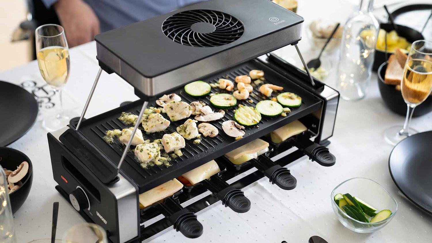 Auf einem Esstisch grillt ein Raclette-Grill Gemüse und Fleischstücke, während in den darunterliegenden Schalen Käse schmilzt. Der Tisch ist mit Champagnergläsern und Schüsseln mit Gemüse gedeckt.