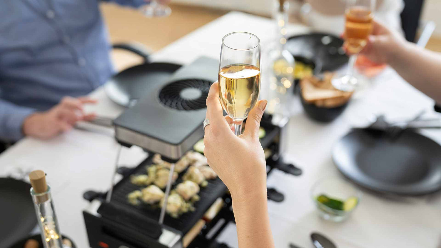 Eine Hand hält ein Glas Sekt hoch, um auf einen Esstisch anzustoßen, der mit Tellern, Getränken und einem Tischgrill gedeckt ist, auf dem Essen zubereitet wird; die Hände anderer Personen sind sichtbar.