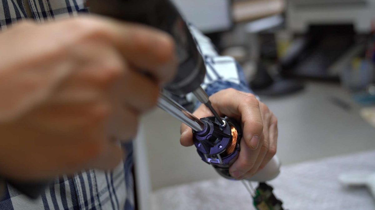 Nahaufnahme einer Person, die mit einer Bohrmaschine ein kleines mechanisches Gerät zusammenbaut oder repariert. Die Hände sind scharf dargestellt, der Hintergrund ist unscharf und zeigt einen Arbeitsbereich in einem Innenbereich.