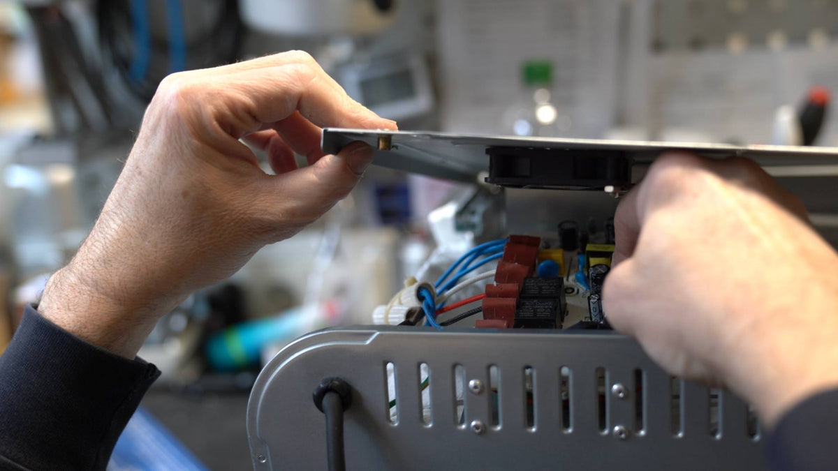 Die Hände einer Person montieren oder reparieren ein elektronisches Gerät und befestigen in einer Werkstattumgebung eine Metallabdeckung an einer Platte mit sichtbaren Kabeln, Anschlüssen und Schaltungskomponenten.