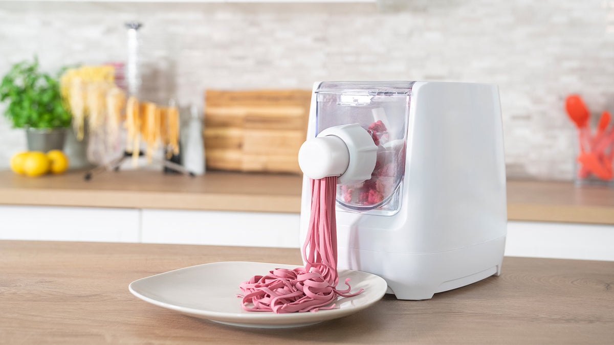 Eine weiße Nudelmaschine auf einer Küchentheke presst rosa Nudeln auf einen Teller, im Hintergrund sind Küchenutensilien, Blattgemüse und trocknende gelbe Nudeln zu sehen.
