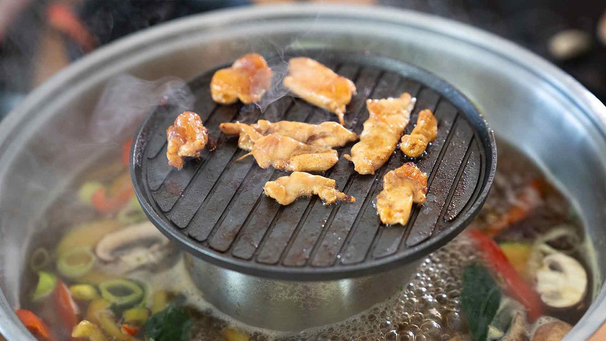 Auf einem runden Metallgrill wird geschnittenes Fleisch über einem Topf mit kochender Suppe und Gemüse gegrillt. So entsteht eine Kombination aus Eintopf und Barbecue. Dampf steigt vom Essen auf und unterstreicht den Garvorgang.