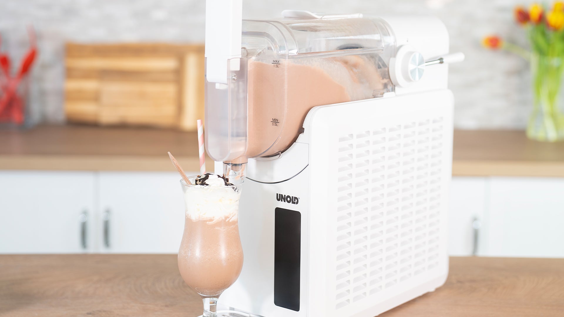 Eine weiße Arbeitsplattenmaschine gibt einen Schokoladenmilchshake in ein hohes Glas mit Schlagsahne, Schokoladensirup und einem Strohhalm aus. Der Küchenhintergrund besteht aus einem Schneidebrett und einer Blumenvase.