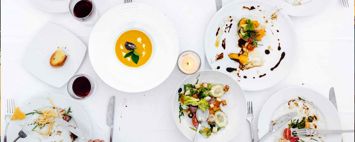 Draufsicht auf einen Esstisch mit Tellern mit teilweise gegessenem Essen, Suppe, Salat, Gemüse, Weingläsern, einer Kerze und Brotstücken auf einer weißen Tischdecke.