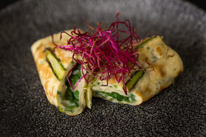 Das Unold Raclette Multi 4 in 1 inklusive Raclette Buch wird auf einem stilvollen dunklen Teller serviert, begleitet von grünem Gemüse und leuchtend violetten Microgreens für eine frische, farbenfrohe Präsentation.