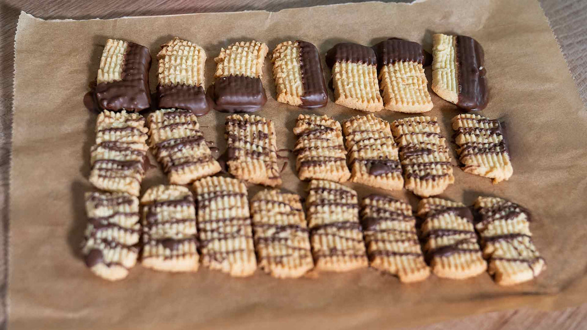 Eine Ladung rechteckiger Butterkekse auf braunem Pergamentpapier, einige zur Hälfte in Schokolade getaucht und andere mit Schokolade beträufelt, in ordentlichen Reihen angeordnet.