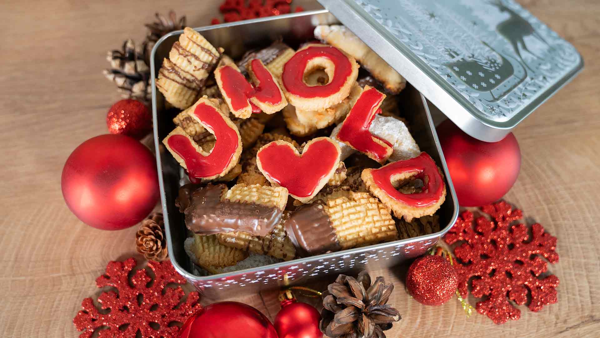 Eine festliche Dose gefüllt mit verschiedenen Keksen, einige davon mit rotem Zuckerguss, der „UNO <3 MOL“ buchstabiert. Die Dose ist umgeben von roten Ornamenten, Tannenzapfen und roten Glitzerschneeflocken auf einer Holzoberfläche.