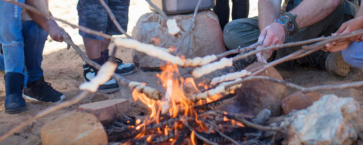 Mehrere Personen sitzen um ein Lagerfeuer und rösten Teig auf Stöcken über den Flammen. Nur ihre Hände und Beine sind zu sehen. Das Feuer ist von Felsen und Sandboden umgeben.