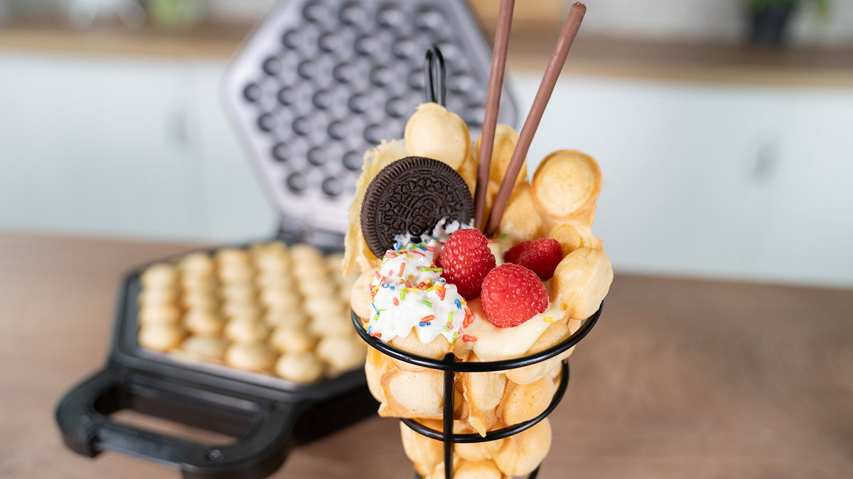 Eine Bubble-Waffel gefüllt mit Schlagsahne, Himbeeren, Streuseln, einem Oreo-Keks und zwei Schokosticks, präsentiert in einem Metallhalter mit einem Waffeleisen und weiteren Waffeln im Hintergrund.