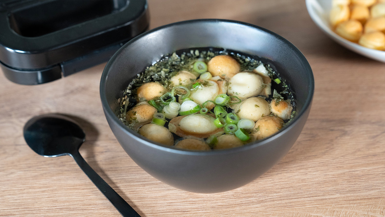 Eine schwarze Schüssel mit Suppe, die kleine runde Knödel, gehackte Frühlingszwiebeln und Kräuter enthält, steht auf einem Holztisch neben einem schwarzen Löffel und einem Teller mit weiteren Knödeln.