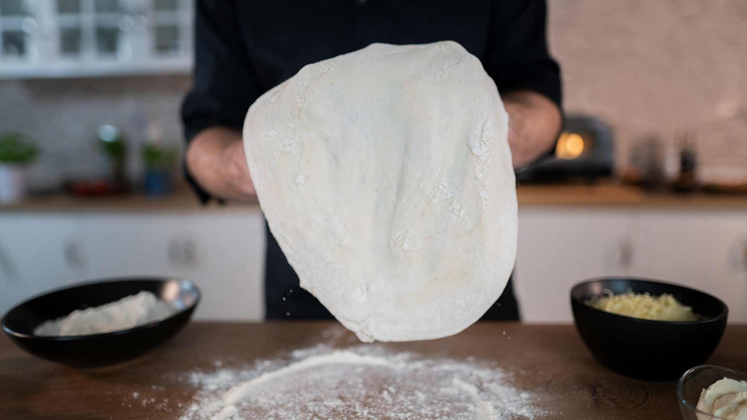 Eine Person hält einen ausgerollten, bemehlten Pizzateig über einen mit Mehl bedeckten Holztisch in einer Küche. In der Nähe stehen Schüsseln mit Zutaten.