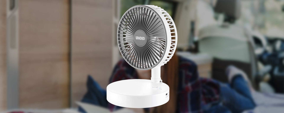 Ein kleiner, weißer, tragbarer elektrischer Ventilator wird im Innenbereich ausgestellt, vor einem unscharfen Hintergrund, der eine gemütliche, ungezwungene Umgebung mit einem Bett und einer Decke zeigt.