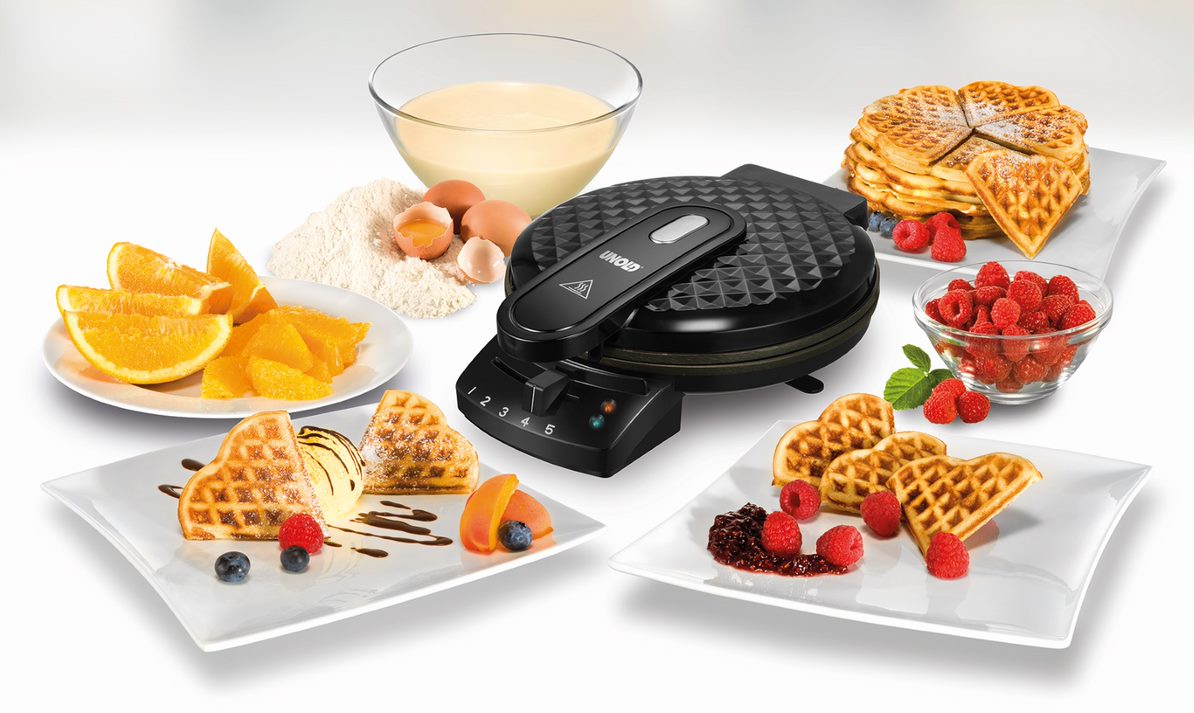 Der Unold Waffelautomat Diamant steht umgeben von Tellern mit Waffeln, garniert mit Früchten, Eiscreme und Sirup, sowie einer Schüssel mit Teig, Eiern, Mehl, Orangenscheiben und einem Stapel einfacher Waffeln.