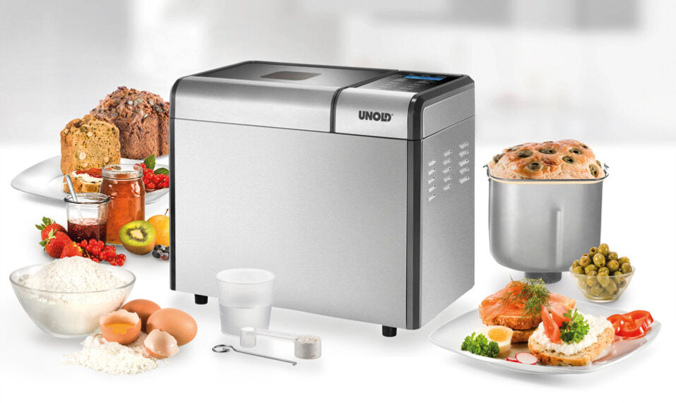Der Brotbackautomat Unold Backmeister® Top Edition steht auf einer weißen Fläche, umgeben von Mehl, Eiern, Butter, Obst, gebackenen Brotlaiben, Tellern mit zubereiteten Speisen und Marmeladengläsern.