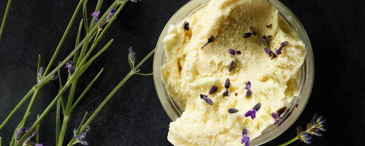 Eine Schale mit cremigem Vanilleeis ist mit kleinen violetten Lavendelblüten garniert. Auf einer dunklen Fläche neben der Schale liegen Zweige frischen Lavendels.