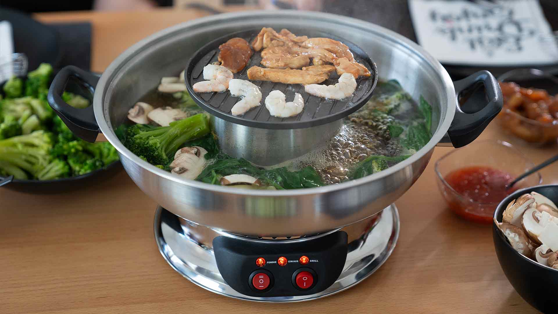 Ein Eintopf mit kochender Brühe enthält Gemüse und Pilze, während darüber auf einem Grill Garnelen, Fleisch und Tofu garen. Teller mit Gemüse und eine Schüssel mit Soße stehen daneben auf dem Tisch.