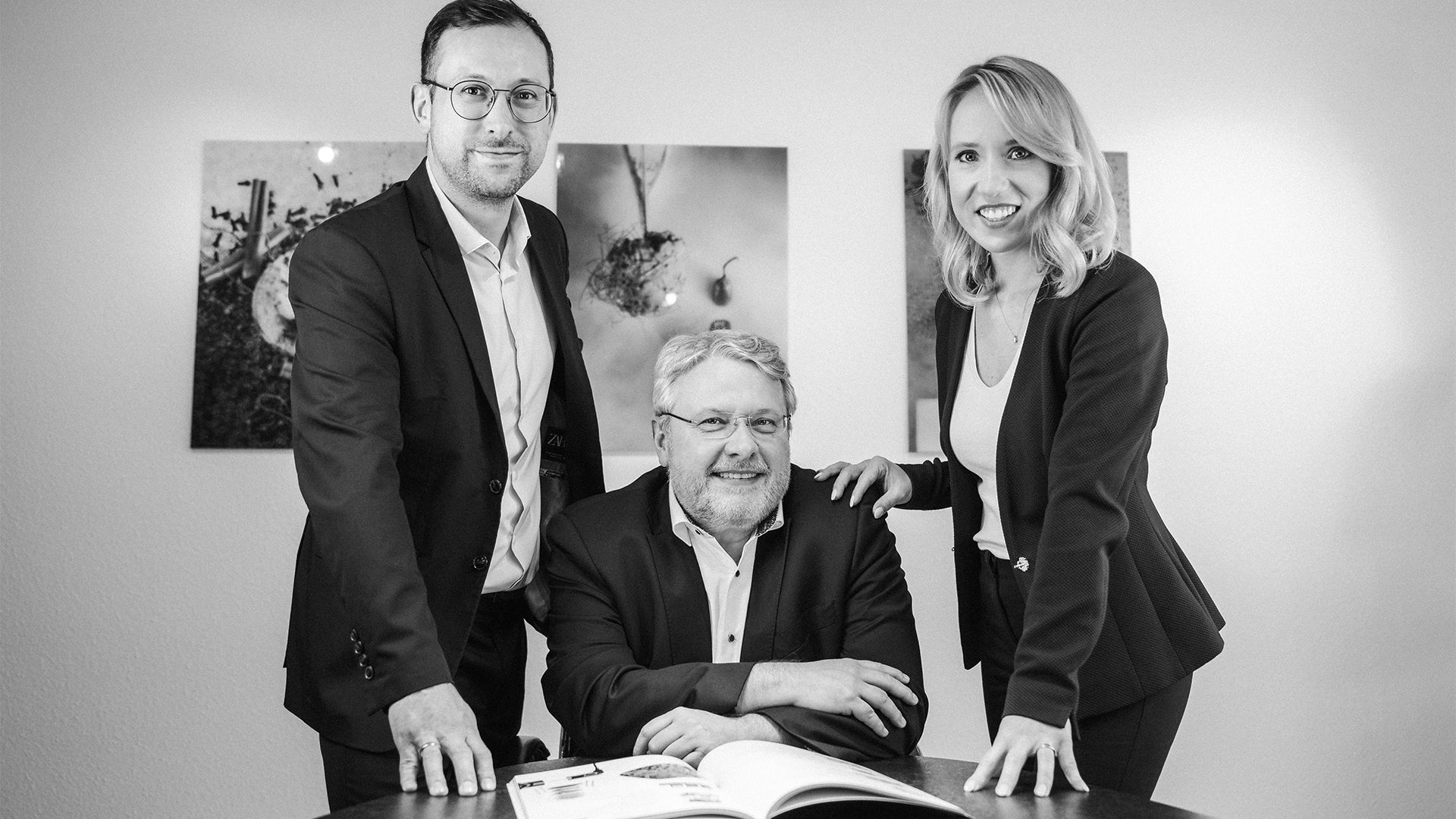 Drei professionell gekleidete Erwachsene – zwei stehend, einer sitzend – posieren gemeinsam mit einem offenen Buch an einem Tisch und lächeln in die Kamera in einem modernen Büro mit gerahmter Kunst an der Wand hinter ihnen.