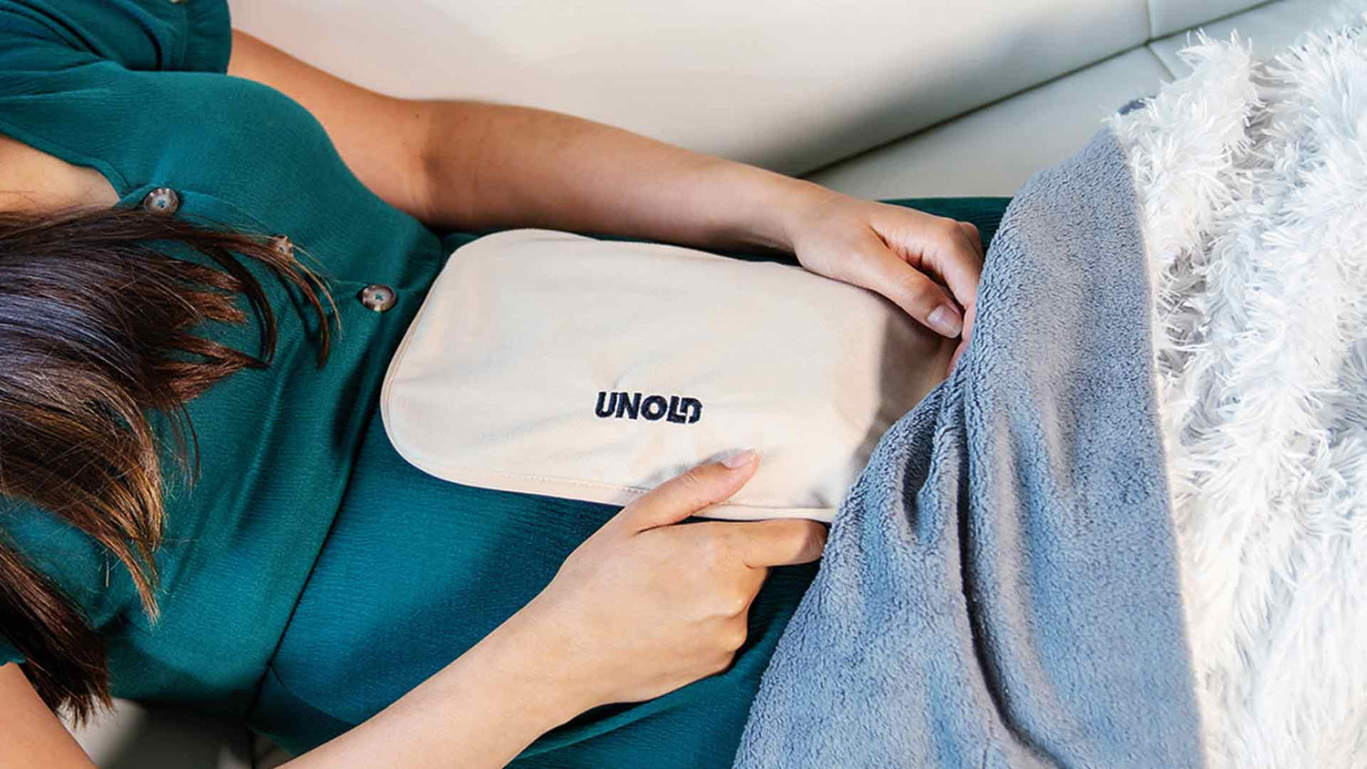 Eine Person in einem grünen Kleid liegt auf einer Couch und hält ein weißes Heizkissen mit der Aufschrift „UNOLD“ auf ihrem Bauch, der teilweise mit einer grauen Decke bedeckt ist.