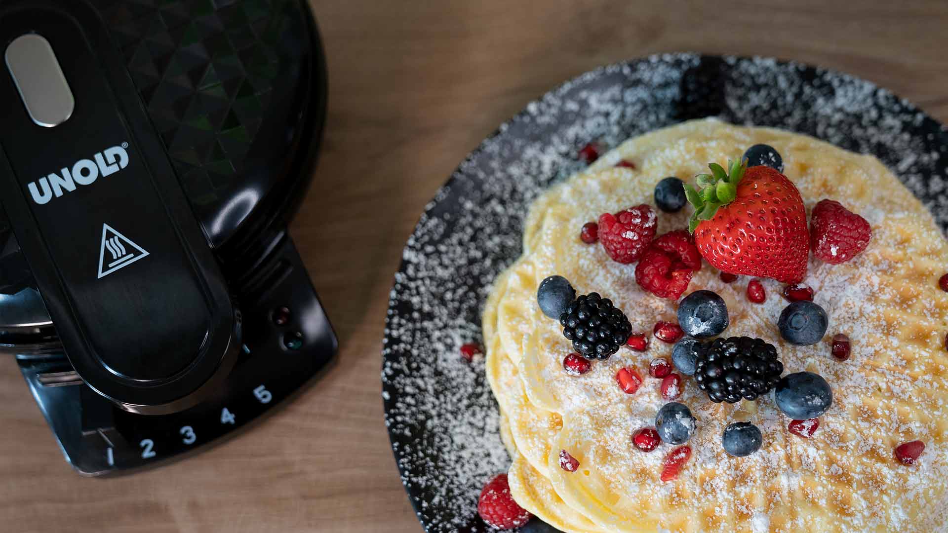 Ein Stapel Pfannkuchen mit Puderzucker, Erdbeeren, Himbeeren, Blaubeeren, Brombeeren und Granatapfelkernen auf einem schwarzen Teller, neben einem schwarzen UNOLD-Waffeleisen auf einer Holzoberfläche.