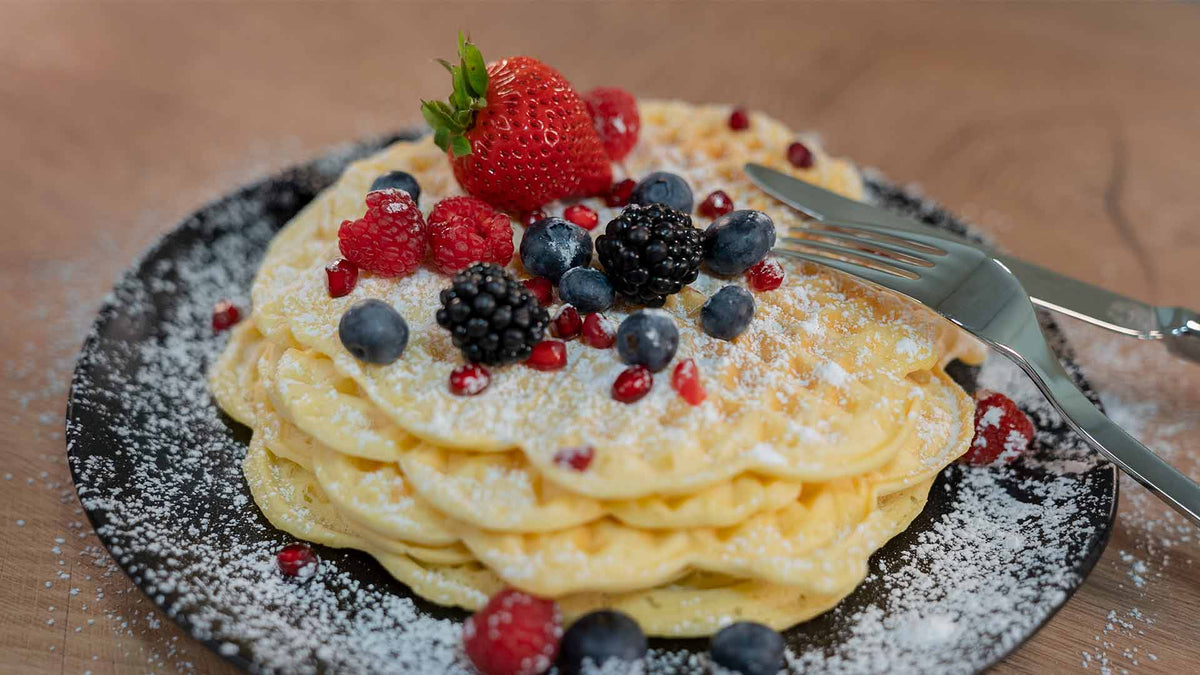 Ein Teller mit Waffeln, garniert mit Erdbeeren, Himbeeren, Brombeeren, Blaubeeren und Granatapfelkernen, bestäubt mit Puderzucker. Gabel und Messer liegen am Rand des Tellers.
