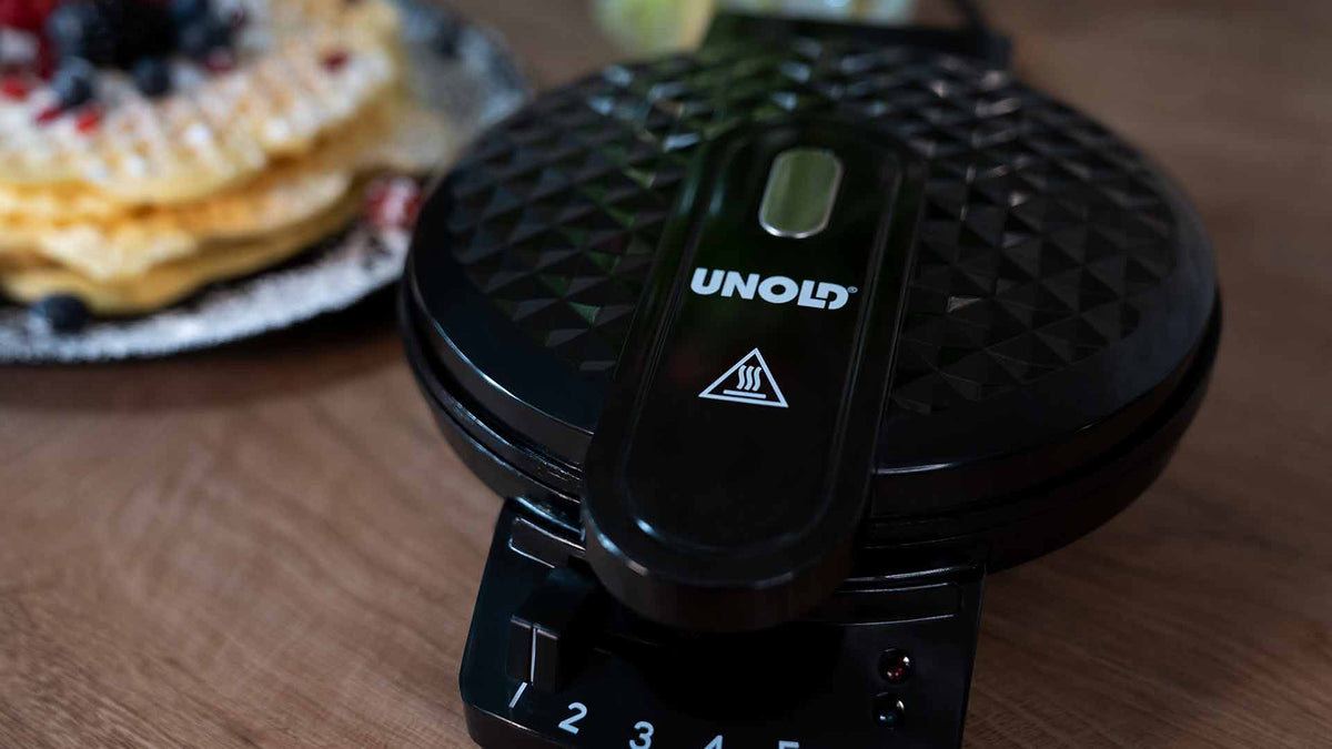 Auf einer Holzoberfläche steht ein schwarzes Waffeleisen von UNOLD, im Hintergrund ist ein Teller mit goldenen Waffeln und Beeren zu sehen.