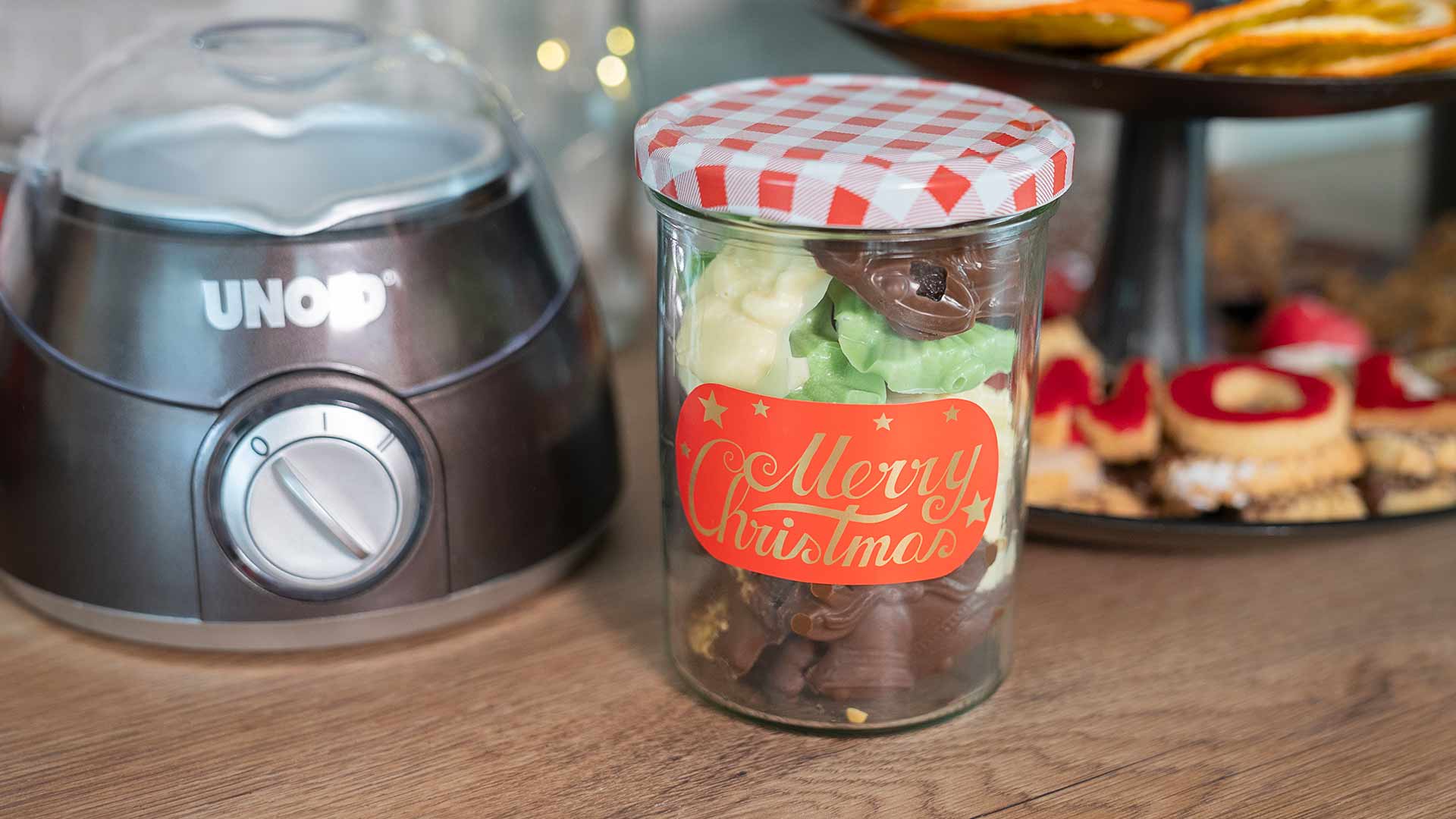 Ein Glas mit der Aufschrift „Frohe Weihnachten“ und einem rot-weiß karierten Deckel ist mit Schokolade gefüllt. Es steht auf einer Holztheke neben einem Küchengerät und einem Tablett mit Weihnachtsplätzchen.