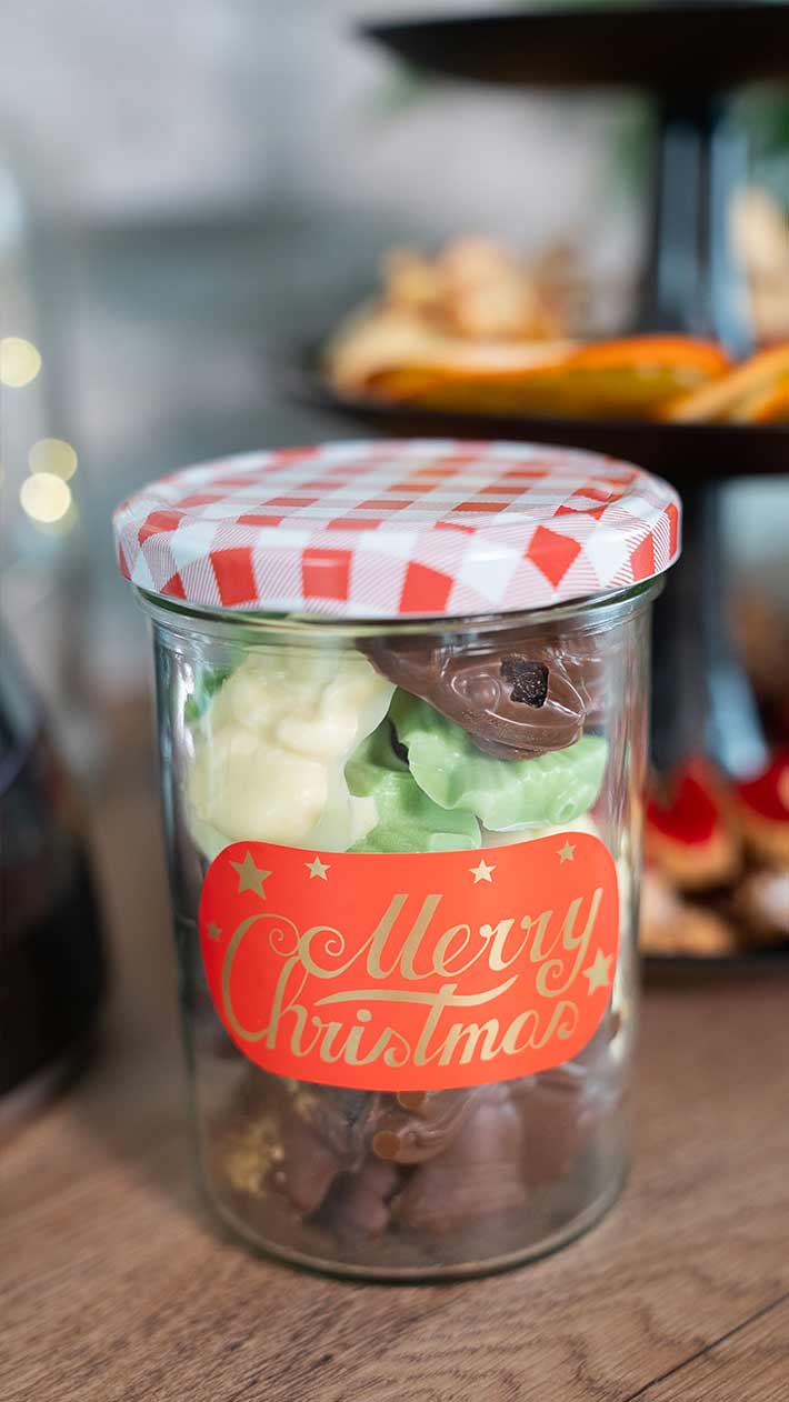 Auf einem Tisch steht ein Glas mit rot-weiß kariertem Deckel, gefüllt mit verschiedenen Pralinen und Bonbons. Das Glas hat ein festliches rotes Etikett mit der Aufschrift „Frohe Weihnachten“.