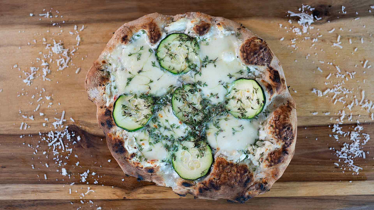 Eine rustikale Pizza mit geschmolzenem Käse, Zucchinischeiben und Kräutern liegt auf einer Holzoberfläche, darüber ist geriebener Käse verstreut.