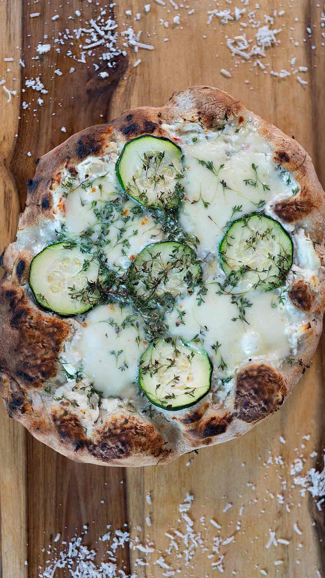 Eine frisch gebackene Pizza mit geschmolzenem Käse, Zucchinischeiben und frischen Kräutern liegt auf einem Holzbrett, über das geriebener Käse verstreut ist.