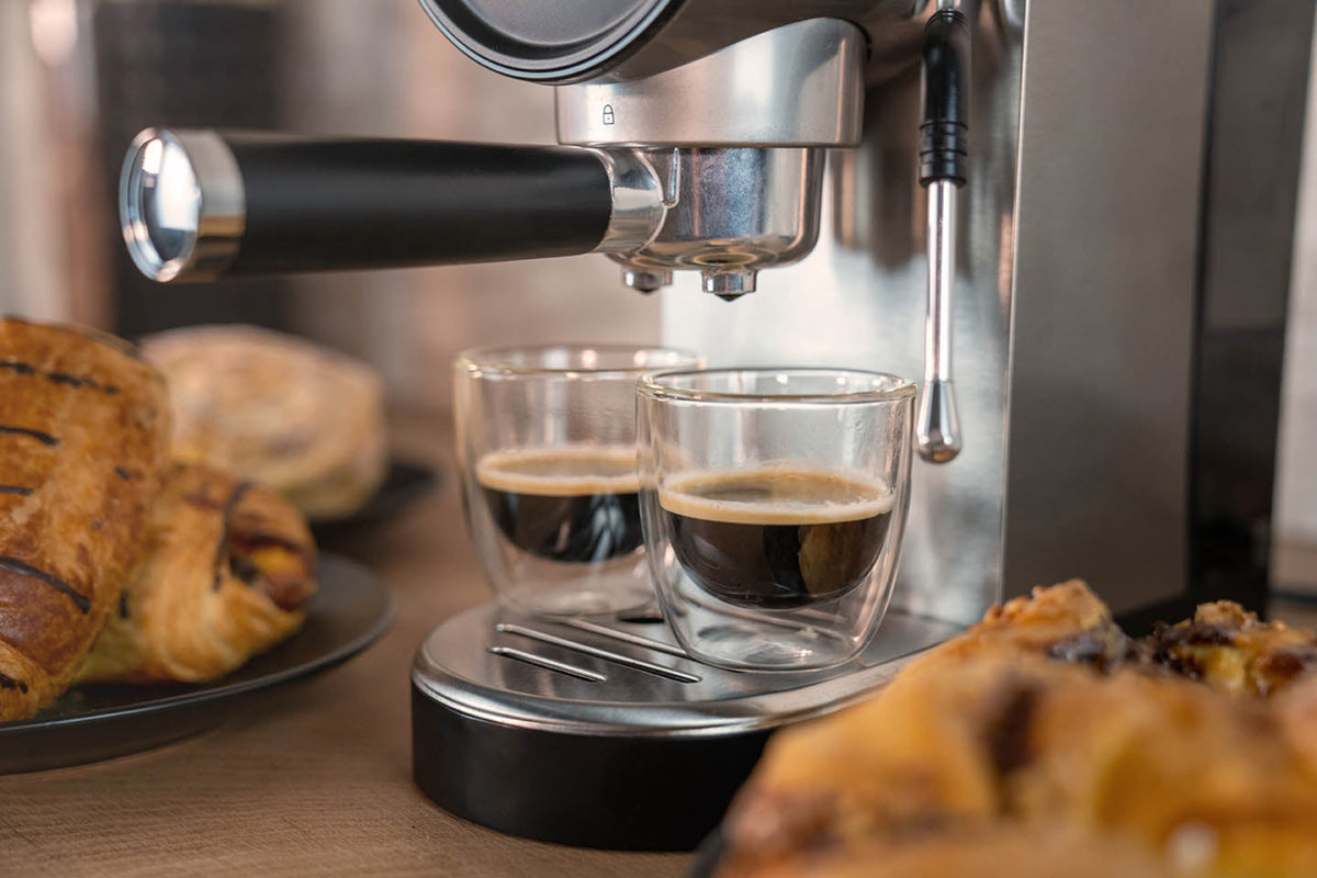 Eine Nahaufnahme der Unold Espressomaschine Piccopresso, die zwei Shots in Glastassen brüht, mit Tellern mit Gebäck, die im Vordergrund und Hintergrund auf einer Holzoberfläche angeordnet sind.