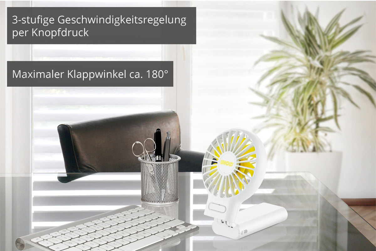 Der Unold HANDVENTILATOR BREEZY FOLD, ein kleiner weißer Tischventilator, steht auf einem Glastisch neben Büromaterial und einer Tastatur. Der deutsche Text hebt die 3-stufige Tastensteuerung und den ca. 180-Grad-Klappwinkel hervor.